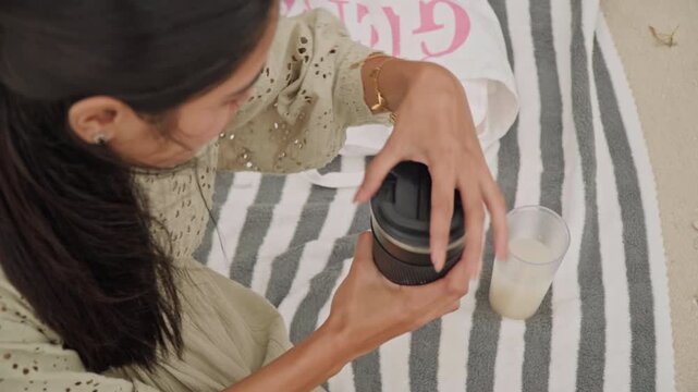 Woman preparing beverage by seaside relaxing setting. Close up of hands opening jar beside glass of milk. Serene beach scene with woman stirring coffee near milk glass and striped blanket
