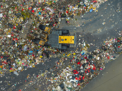 Aerial view of a yellow bulldozer working in a large landfill site surrounded by piles of plastic waste and debris in Jakarta, Indonesia.