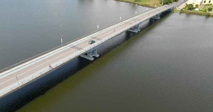 Aerial view of the Sabaudia Bridge, Lazio, Italy. It's a modern low-profile bridge. It features a pedestrian and bike path and a car lane, supported by columns over dark water, with a grassy riverbank