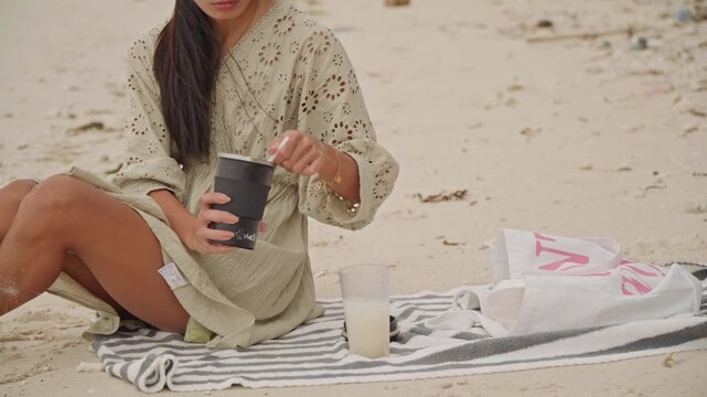 Woman seated on beach towel stirring coffee jar, legs folded on sand, striped blanket and glass of milk beside, relaxed seaside breakfast mood, effortless bohemian style and natural sunlight