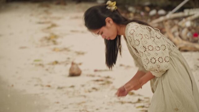 Young asian woman on beach picking shells, wearing lace dress with flower in hair, strolling near driftwood and moored boat, examining finds by shoreline, quiet contemplative mood with warm soft light