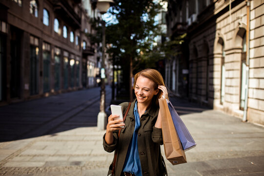 Smiling woman with shopping bags using smartphone on city street