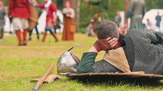 A medieval reenactor lies on the ground, wearing a chainmail shirt and a simple tunic. In the background, various warriors in historical attire engage in a mock battle, showcasing traditional weapons