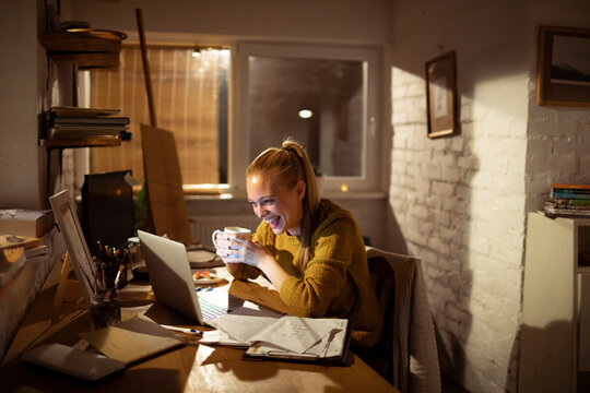 Young woman laughing over laptop with coffee in cozy home office at night