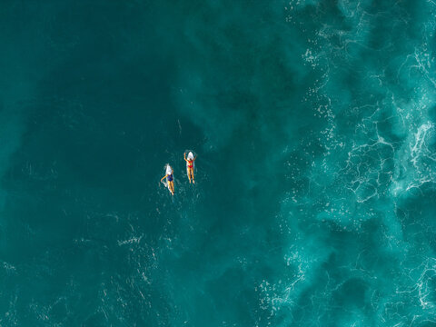Aerial view of two surfers paddling on their boards in the deep turquoise ocean water off the coast of Bali, Indonesia.