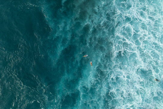 Aerial view of surfers paddling on their boards in the turquoise ocean with white crashing waves in Bali, Indonesia.