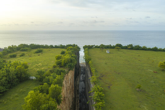 Aerial view of the road cut through the limestone cliffs at Pantai Tanah Barak, leading to the ocean, surrounded by green fields in South Kuta, Bali, Indonesia.