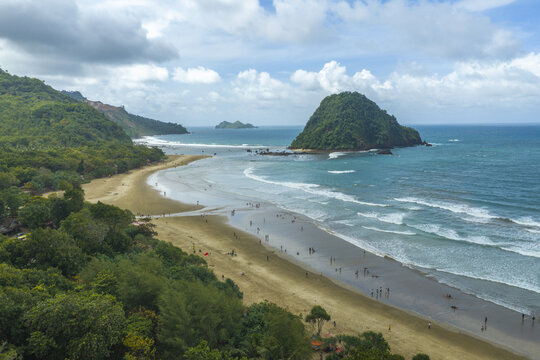 Aerial view of Pantai Pulau Merah with its distinctive red-hued hill, sandy beach, turquoise waves, and lush green forest in Kecamatan Pesanggaran, Jawa Timur, Indonesia.