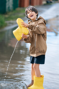 Happy boy pours water from his yellow rainboot into a puddle