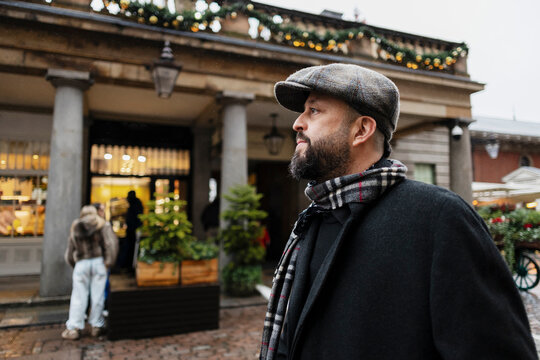 A bearded man wears a flat cap at a festive winter market outdoors