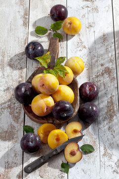 Sliced and whole yellow and black gages (Prunus domestica subsp. italica) and a wooden spoon on white wooden table, studio shot