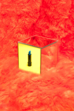 Blue lipstick sits inside a mirrored cube on a red fluffy surface