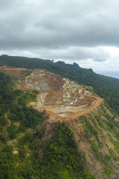 Aerial view of an open-pit gold and copper mine with terraced excavation levels surrounded by dense tropical forest in Kecamatan Pesanggaran, Jawa Timur, Indonesia.