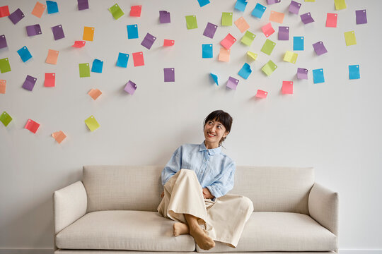 Woman sits on couch looking up at sticky notes in office