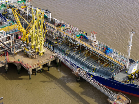 Aerial view of a large tanker ship docked at an industrial terminal with yellow cranes and metal walkways over brown water in the United Kingdom.