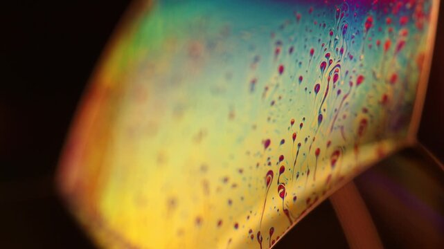 Curved fluid membrane plane with splatter pattern and rainbow light interference. A curved liquid film plane illuminated in low-key macro perspective shows intricate fluid droplets