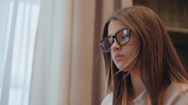Caucasian woman with glasses studying near window soft natural light, long brown hair, serious focused expression, typing on laptop, reading notes, freelance writer and researcher roles, quiet modern