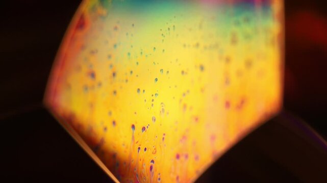 Glowing golden fluid film facet with speckled droplets and cyan gradient. Crisp specular rim lines outline a geometric liquid film facet in low-key macro perspective.