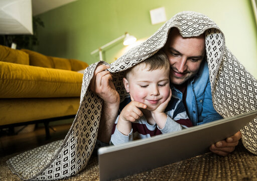 Father and son watch a tablet under a blanket at home