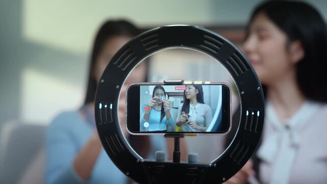 Beauty bloggers influencers live streaming makeup tutorial using ring light and smartphone, video capture in home studio as two women share skincare product review, smile, engage viewers