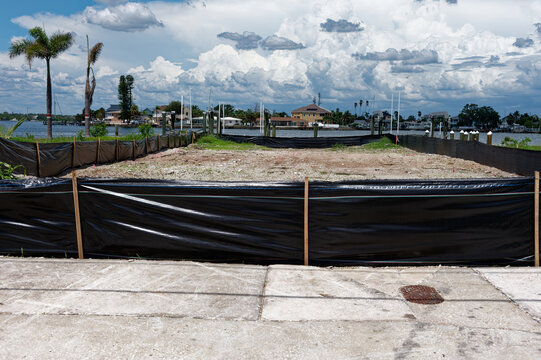 Empty waterfront lot bordered by black silt fencing and stakes, ready for construction, set against dramatic storm clouds and a calm bay with palm trees and residential docks.
