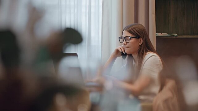 Caucasian woman contemplating at laptop, resting chin on hand, reviewing project documents, soft natural light through sheer curtains, glassware and jacket on chair, thoughtful expression.