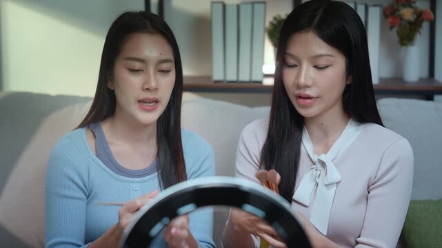 Beauty bloggers influencers live streaming makeup tutorial ring light brush guide as two young women on sofa share glow skincare tips, engaging viewers in home studio with smartphone warm light