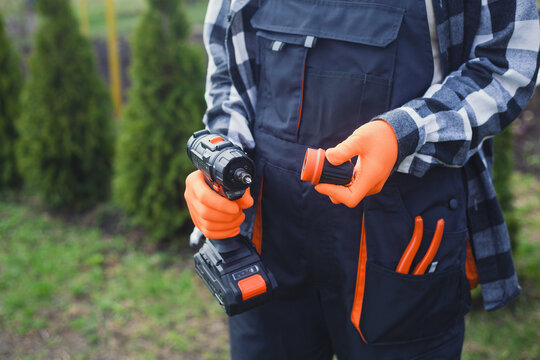 person holding power drill outdoors wearing orange gloves and black jacket for home improvement project in garden setting close-up practical and serene mood natural light clean background.