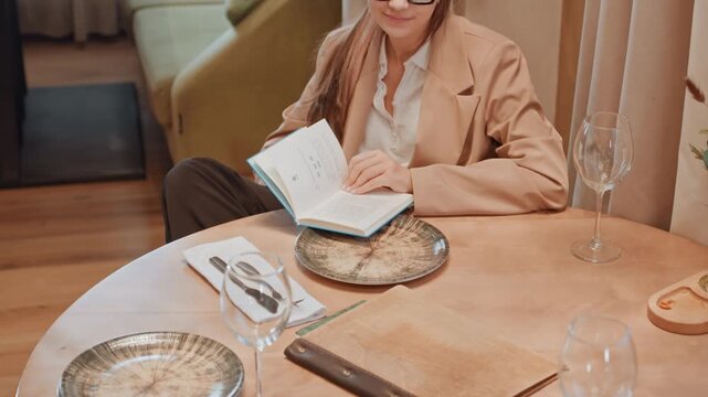 Caucasian woman reading beside set table. Executive consultant reviewing notes during quiet lunch pause, empty chairs around round wooden table, polished plates and glassware, smartphone on table,