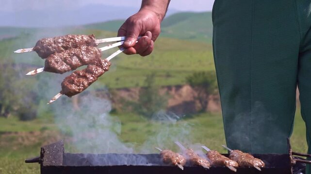 Man holding three skewers of juicy lula kebab shashlik over a smoking mangal grill in a scenic green mountain landscape, horizontal video.