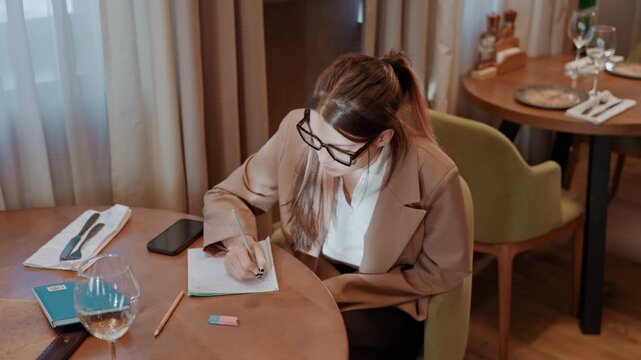 Caucasian woman checking invoices at table calculating expenses with notebook and phone pens scattered receipt visible focused entrepreneur reconciling accounts in quiet bistro warm light