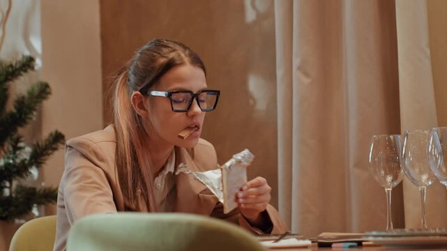 Businesswoman at table eating wrapped snack, pausing between notes and laptop, glasses perched, warm restaurant interior with holiday tree, quick work break with satisfied expression, candid editorial