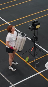 Female coach loading badminton shuttle launcher on indoor court assistant watches while coach lifts cylindrical shuttle feeder onto tripod machine, adjusts height and calibration, aligns camera