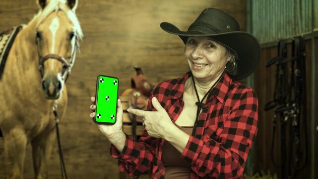 Senior cowgirl presenting a smartphone with green screen beside a palomino horse in a stable. Ideal for app mockups, marketing and technology. 4K.