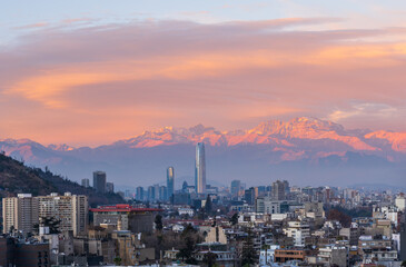 Fototapeta premium Panoramic view of Santiago de Chile city skyline at sunset with the snow capped Andes mountains under a colorful orange and pink sky