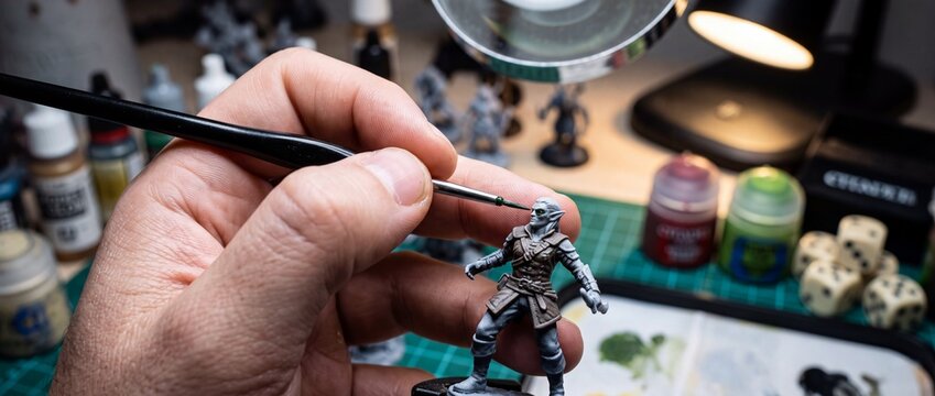 Artist hand painting intricate details on miniature fantasy elf figurine using fine brush under magnifying lamp in creative studio