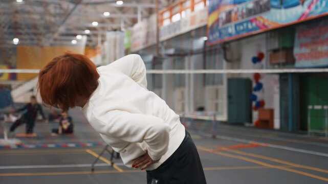 Redhead white woman on badminton court bending backward with hands on lower back during warmup, indoor gymnasium with net and tripod camera, wearing white hoodie and black shorts, focus on posture