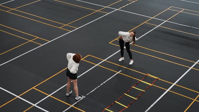 Two players practicing badminton on asphalt, coach guiding footwork beside agility ladder, aerial view of marked training grid showing racket swings, focused warmup and precision step drills