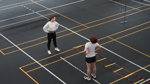 White coach conducting badminton performance analysis with stopwatch timing footwork drills, player executing ladder steps, measurement markers, focused evaluation, indoor court setting
