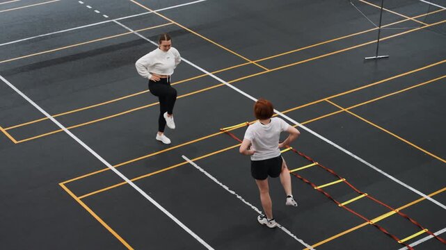 Badminton court players doing ladder drills, overhead view of coach guiding athlete through rapid footwork on marked indoor court with agility ladder, focused tempo, coordinated warmup routine
