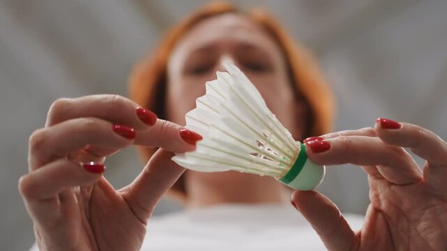 Closeup shuttlecock preparation by white woman, redhaired player adjusts feather skirt with painted red nails, slow careful grip, indoor court roof blurred overhead, prematch ritual, tactile