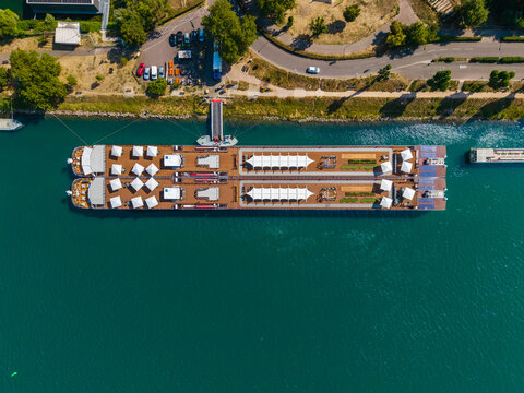 Aerial view of a river cruise ship docked at a pier on the Rhine river with white umbrellas and deck seating in Vogelgrun, Grand Est, France.