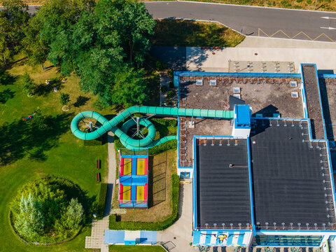 Aerial view of a green water slide winding over a building roof and adjacent park area with trampolines in Vogelgrun, Grand Est, France.