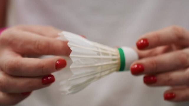 Handson shuttlecock inspection by white woman, redhaired hands with red manicure adjust feather alignment and cork base, methodical tutorial vibe, precision check for training session, indoor court
