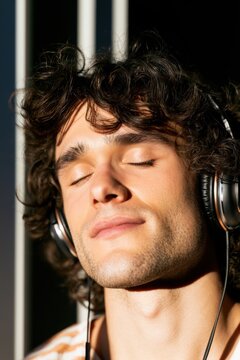 Young man with curly hair wearing over-ear headphones relaxes with eyes closed, illuminated by sunlight streaming through a window in a modern indoor setting