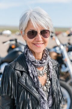 Stylish mature woman with short silver hair wearing sunglasses and a fringed leather jacket, posing outdoors with motorcycles in the background