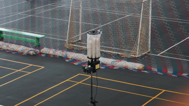 Empty indoor badminton court with launcher shuttlecock machine on tripod near net green bench along sideline yellow court lines colorful barrier behind net single shuttle on floor quiet gym atmosphere