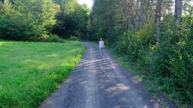 Young brunette woman walks along gravel road past lush green bushes and blooming summer wildflowers. Peaceful rural landscape of Croatia offers relaxation and slow living vibes.