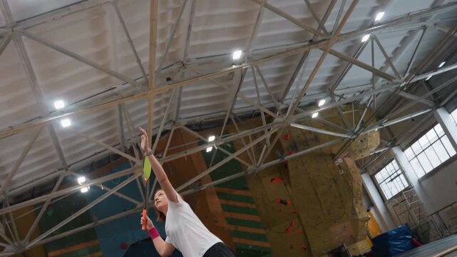 Overhead badminton smash by white player on indoor court, explosive followthrough and elevated jump, concentrated expression and wristband accent, gym roof lights and climbing wall background, dynamic