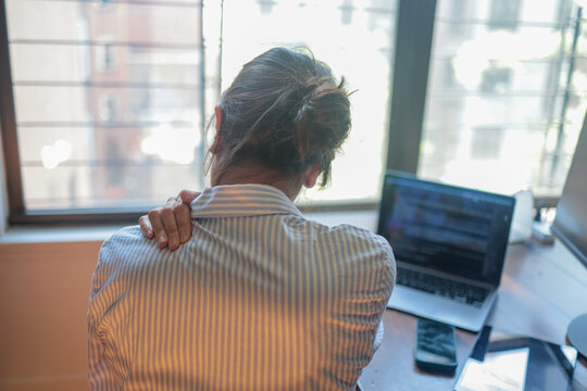 Woman experiencing shoulder and neck pain while working from home on a laptop, feeling discomfort, fatigue, and stress from prolonged bad posture and sedentary lifestyle